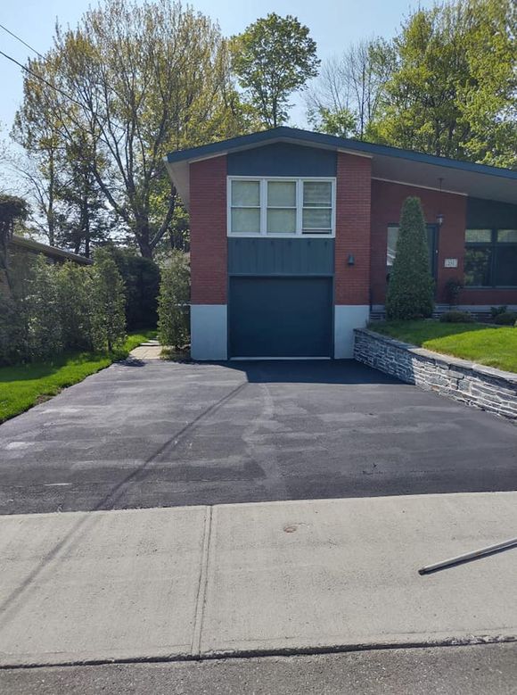 Une maison moderne en briques avec une porte de garage et une allée bleu foncé, entourée de verdure.