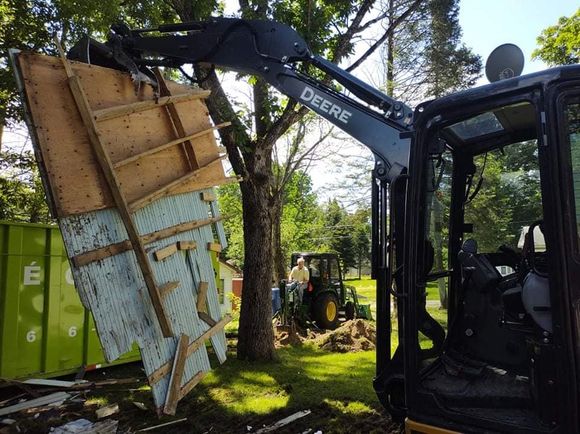 Une pelle mécanique John Deere soulève une partie d'une structure en ruine. Des hommes travaillent à l'arrière-plan. 