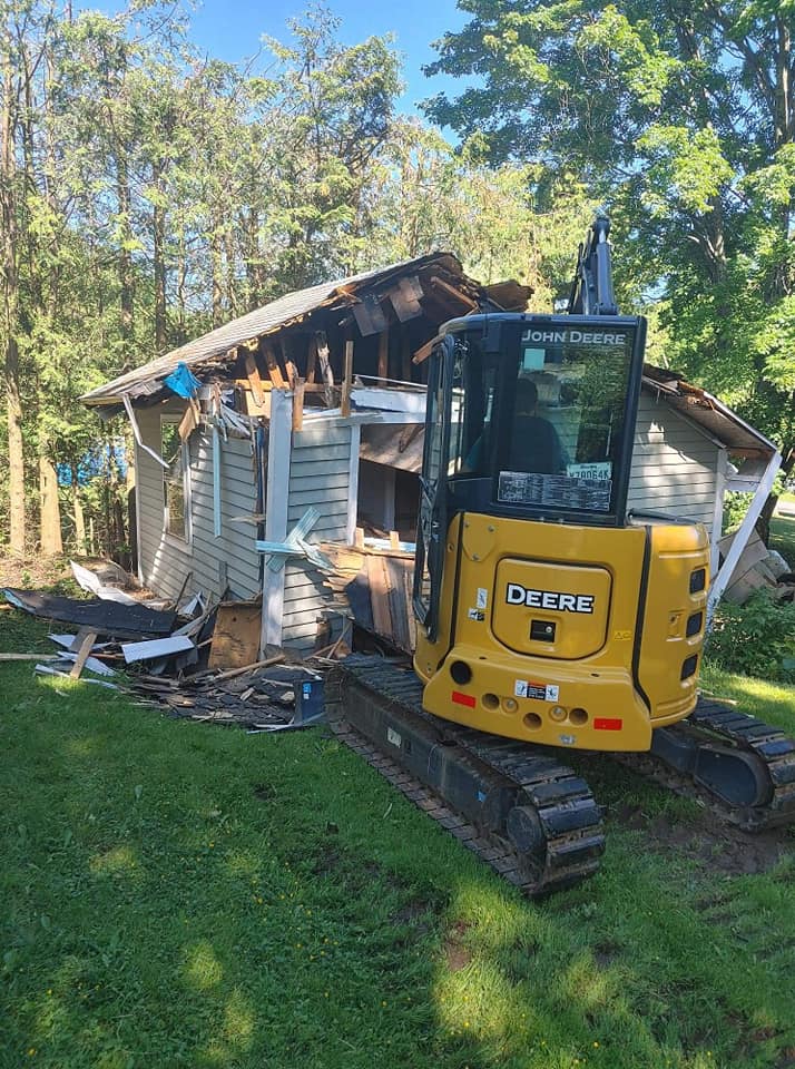 Excavatrice jaune devant une petite maison en démolition dans un boisé.