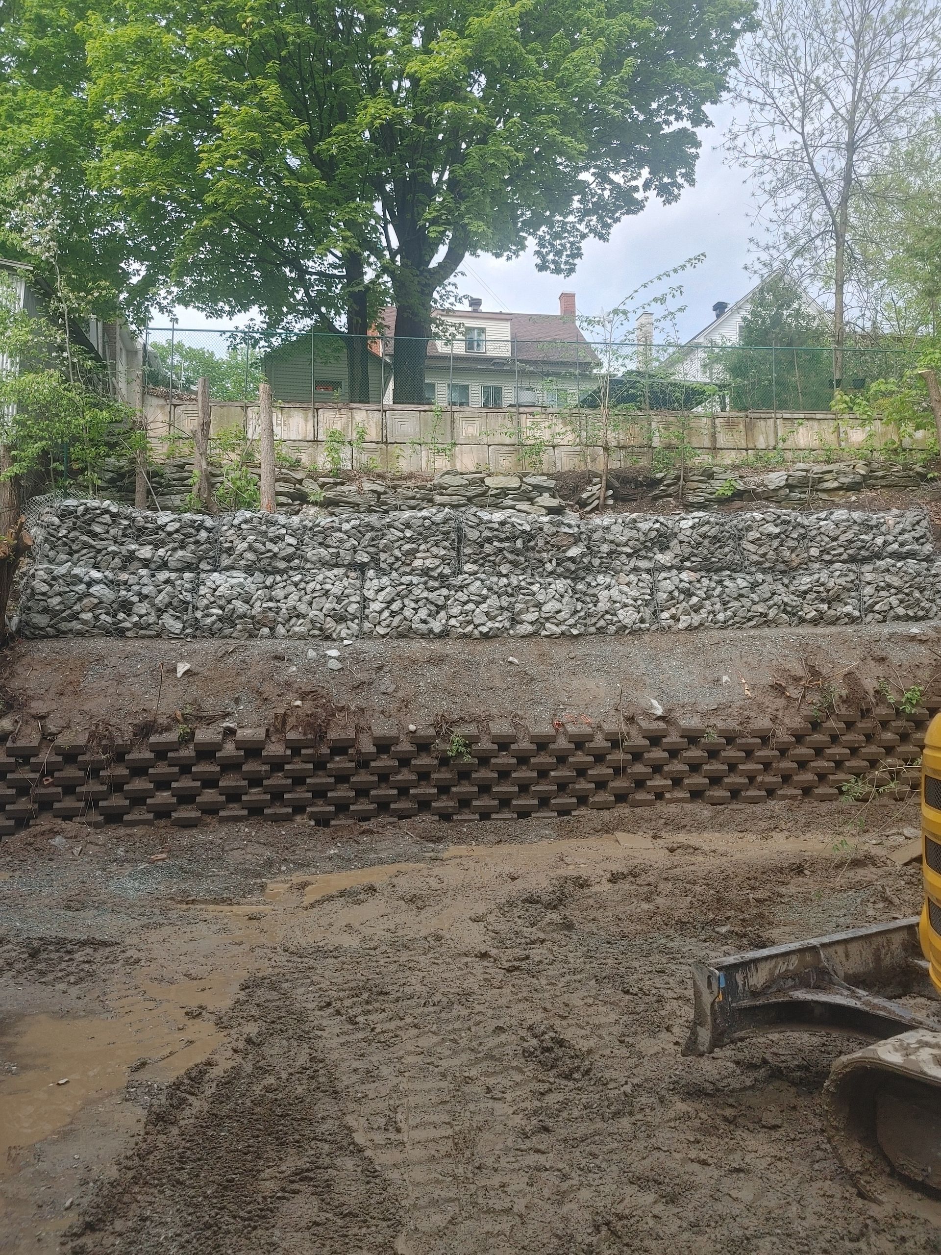 Mur de soutènement en construction : Gabions remplis de roches, de terre et de couches de terre compactée.