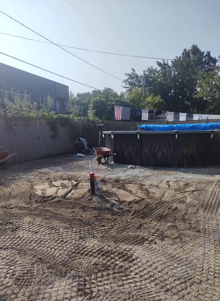 Cour arrière avec piscine hors sol en construction, terrain en terre, brouette et arroseur automatique.