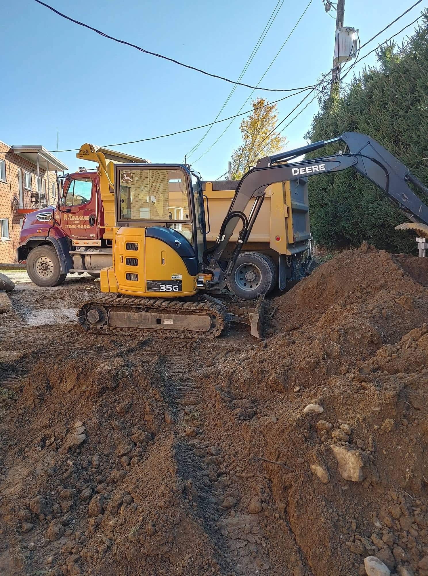 Petite excavatrice jaune chargeant de la terre dans un camion benne sur un chantier résidentiel.