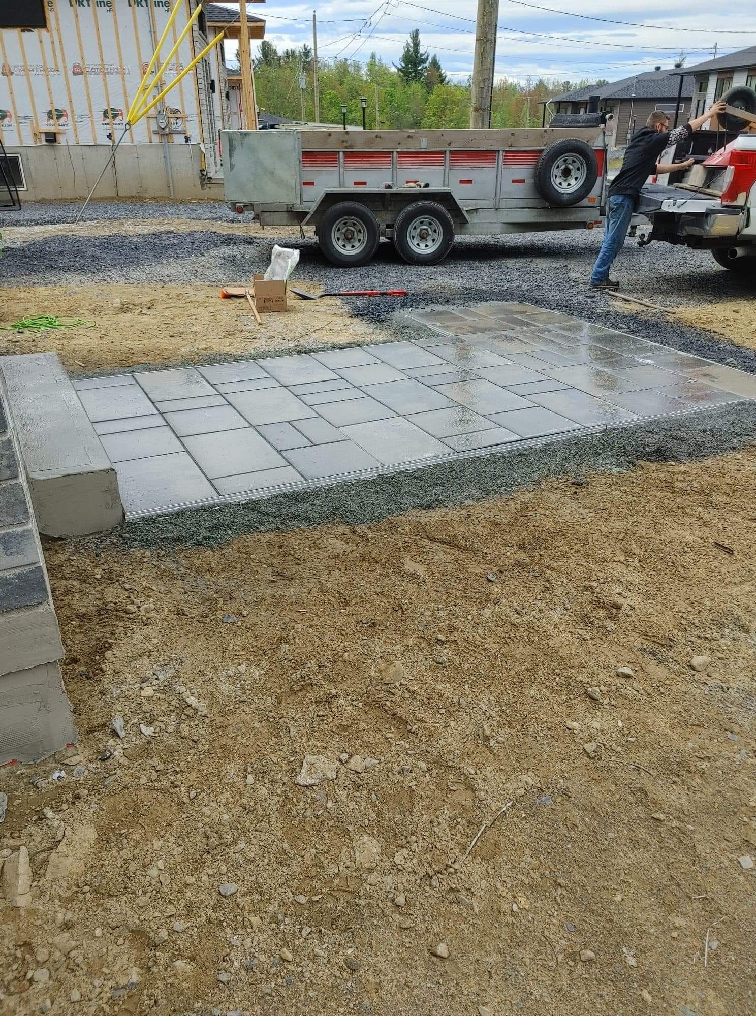 Installation de dalles de b&eacute;ton grises sur un terrain en chantier, avec des camions et des ouvriers en arri&egrave;re-plan.