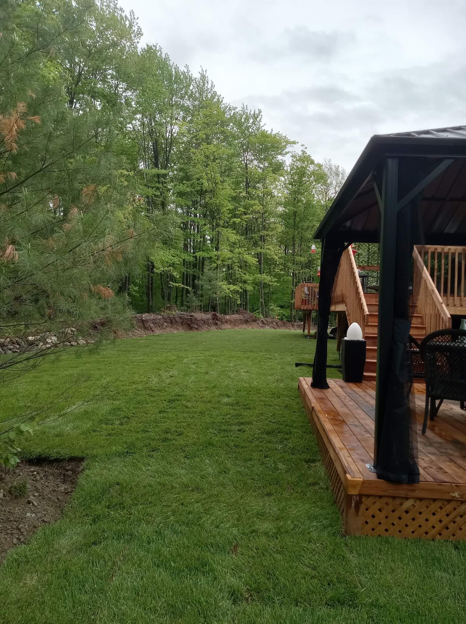 Terrasse en bois avec pergola noire surplombant une pelouse bien entretenue bordée d'arbres.