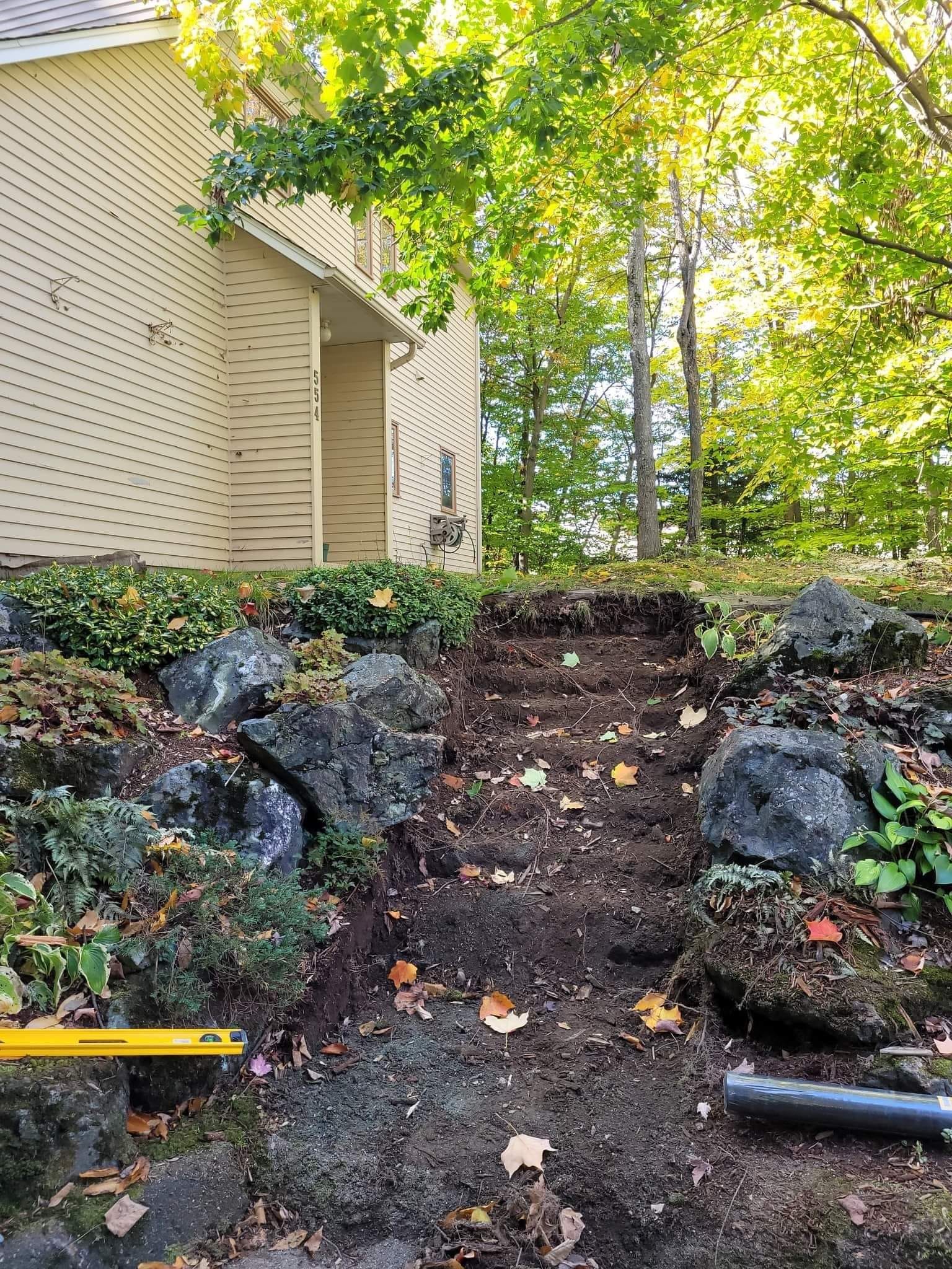 Tranchée excavée devant une maison avec roches de bordure et outils de construction visibles.
