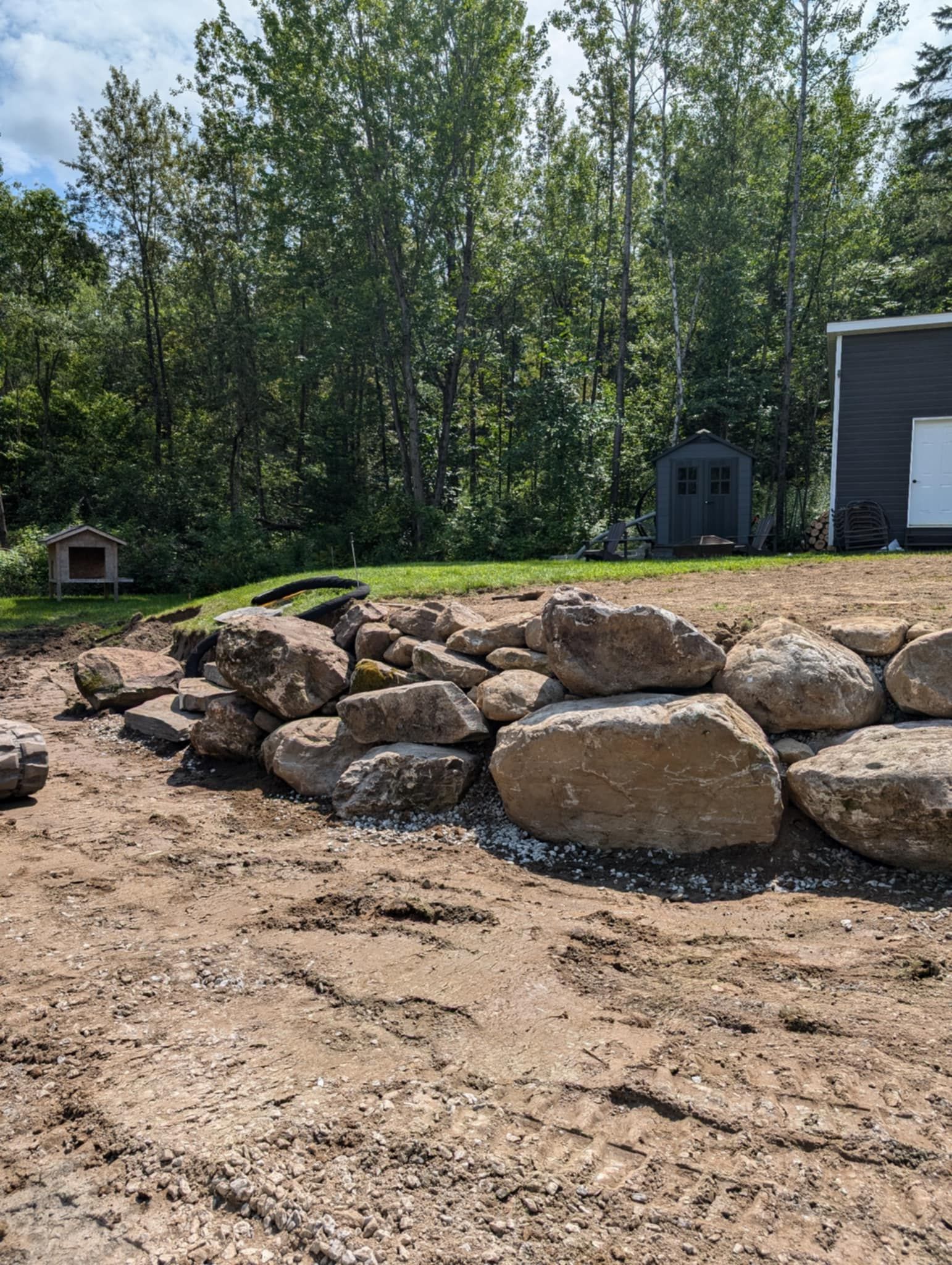 Gros rochers empil&eacute;s formant une structure de retenue sur un terrain nu avec b&acirc;timents et arbres en arri&egrave;re-plan.