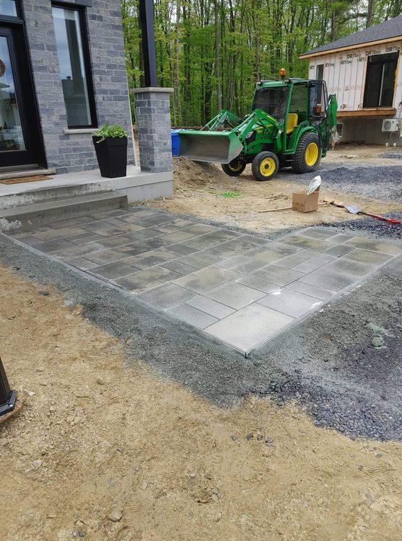 Une terrasse en construction à côté d'une maison, avec un petit tracteur et des bordures en gravier.