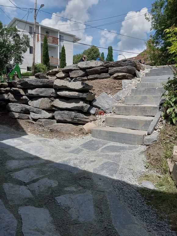 Mur de soutènement en pierre et escalier menant au sommet d'une colline. Un chemin est délimité par du gravier et des pavés.