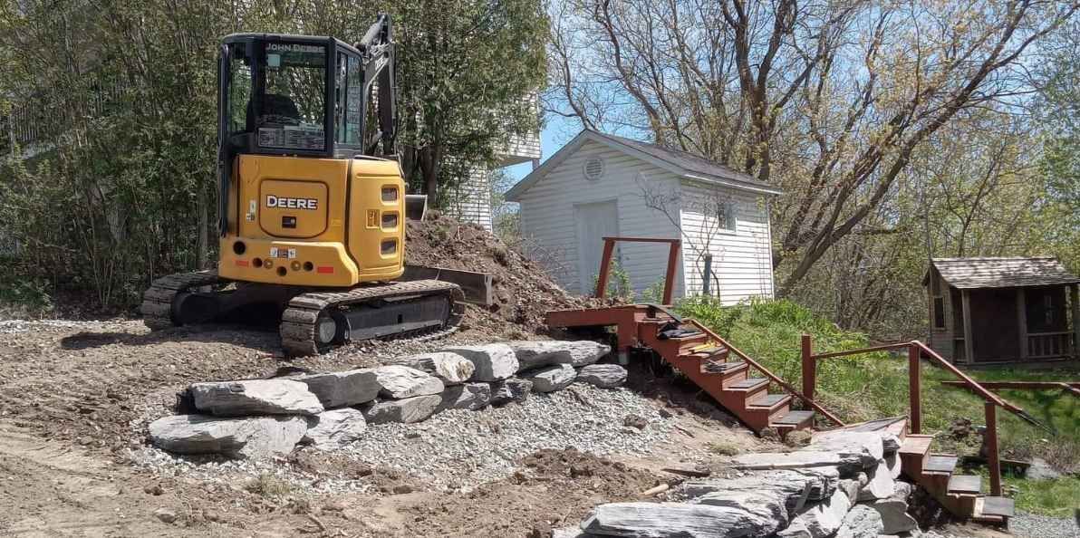 Une pelleteuse jaune et noire se tient sur un terrain accident&eacute; pr&egrave;s d'une maison blanche entour&eacute;e d'arbres nus.