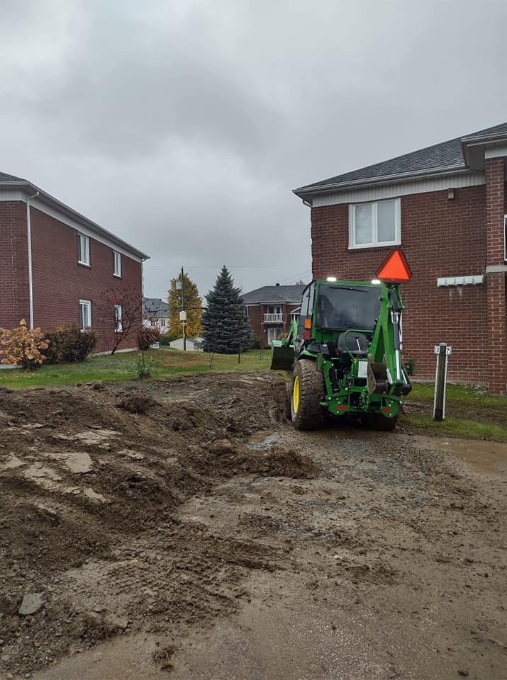 Un tracteur vert et jaune est gar&eacute; devant des maisons en brique rouge avec un terrain de terre fra&icirc;chement excav&eacute;.