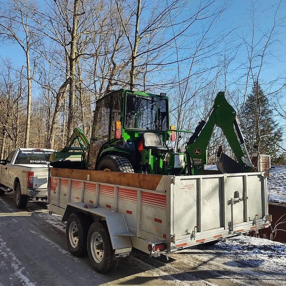 Tracteur vert avec pelle r&eacute;tro charg&eacute; sur une remorque blanche tract&eacute;e par un camion l&eacute;ger.