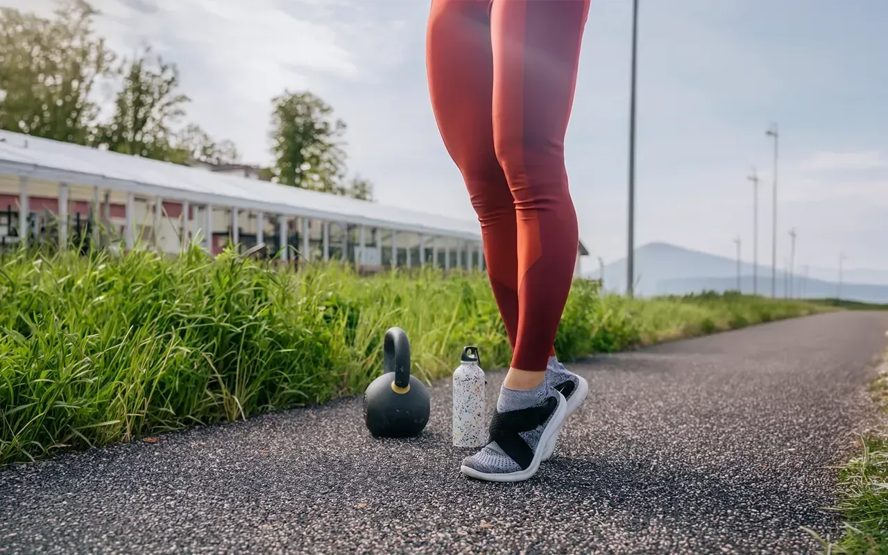 Runner’s legs beside kettlebells on a path, with grass and buildings in the background