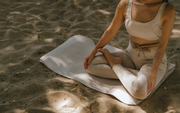 Person seated on a white mat in dappled sunlight, wearing light clothes and meditating outdoors.