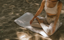 Person sitting cross-legged on a white mat on a sandy floor, wearing a beige tank top and light pants.