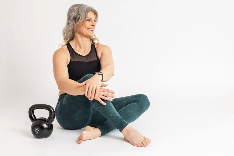 Woman sitting on the floor beside a kettlebell, stretching her legs in a bright studio.