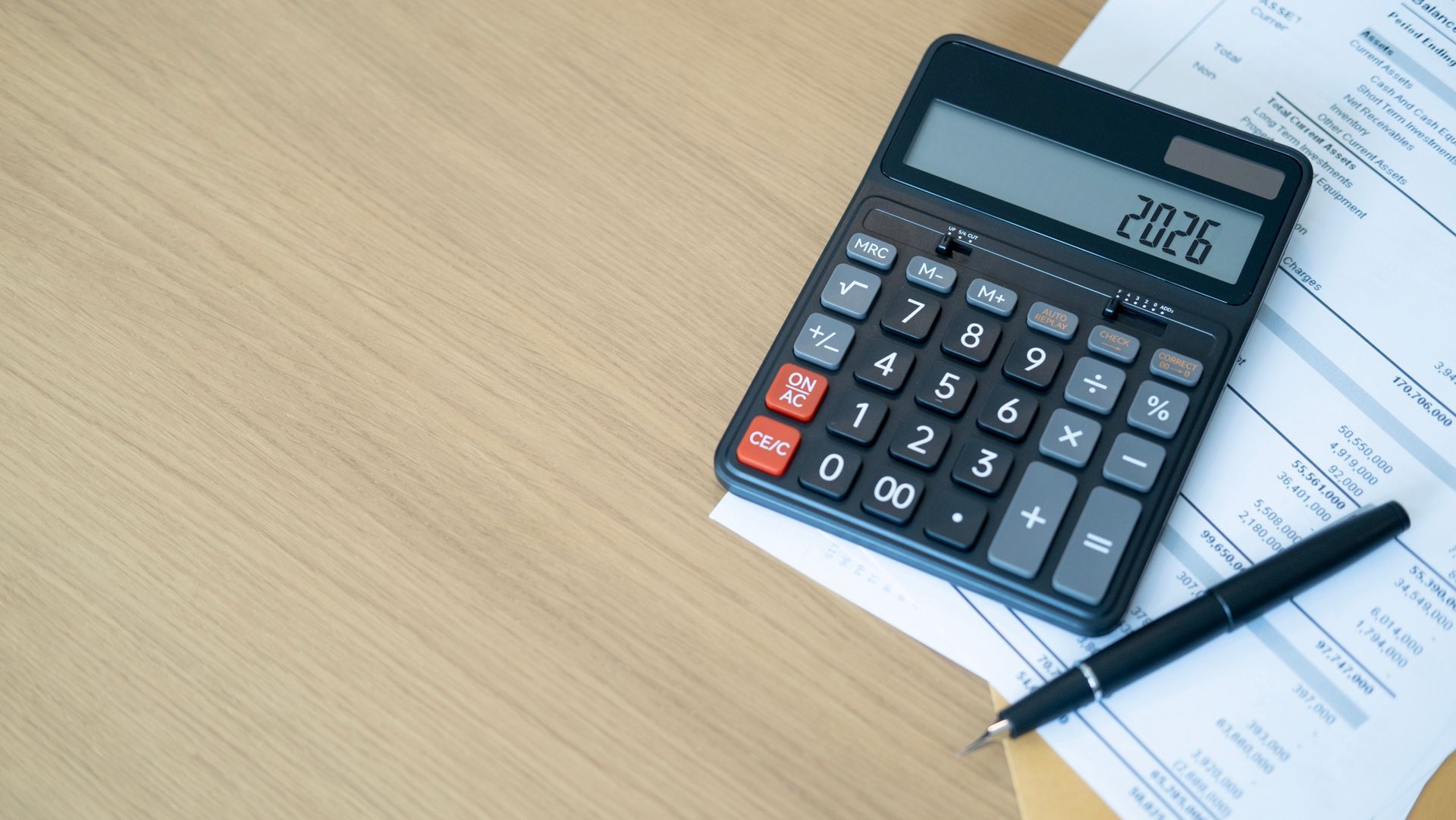 Calculator with 2026 displayed, resting on papers with a pen, on a light wood surface. Calculator with 2026 displayed, resting on papers with a pen, on a light wood surface.