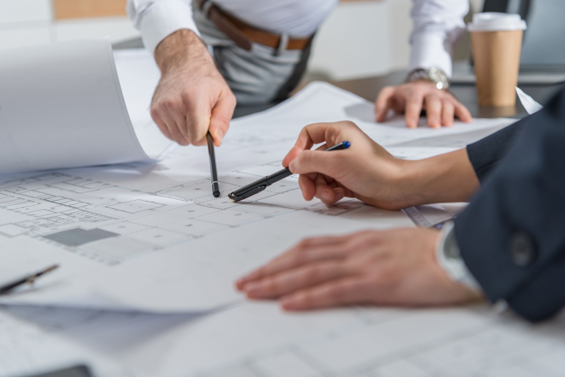 Hands pointing at architectural plans on a table with a laptop and coffee cup nearby Hands pointing at architectural plans on a table with a laptop and coffee cup nearby