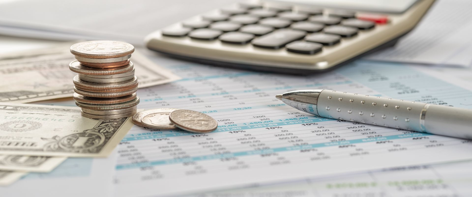 A stack of coins, paper money, a calculator, and a metallic pen arranged on a document with financial data tables. A stack of coins, paper money, a calculator, and a metallic pen arranged on a document with financial data tables.