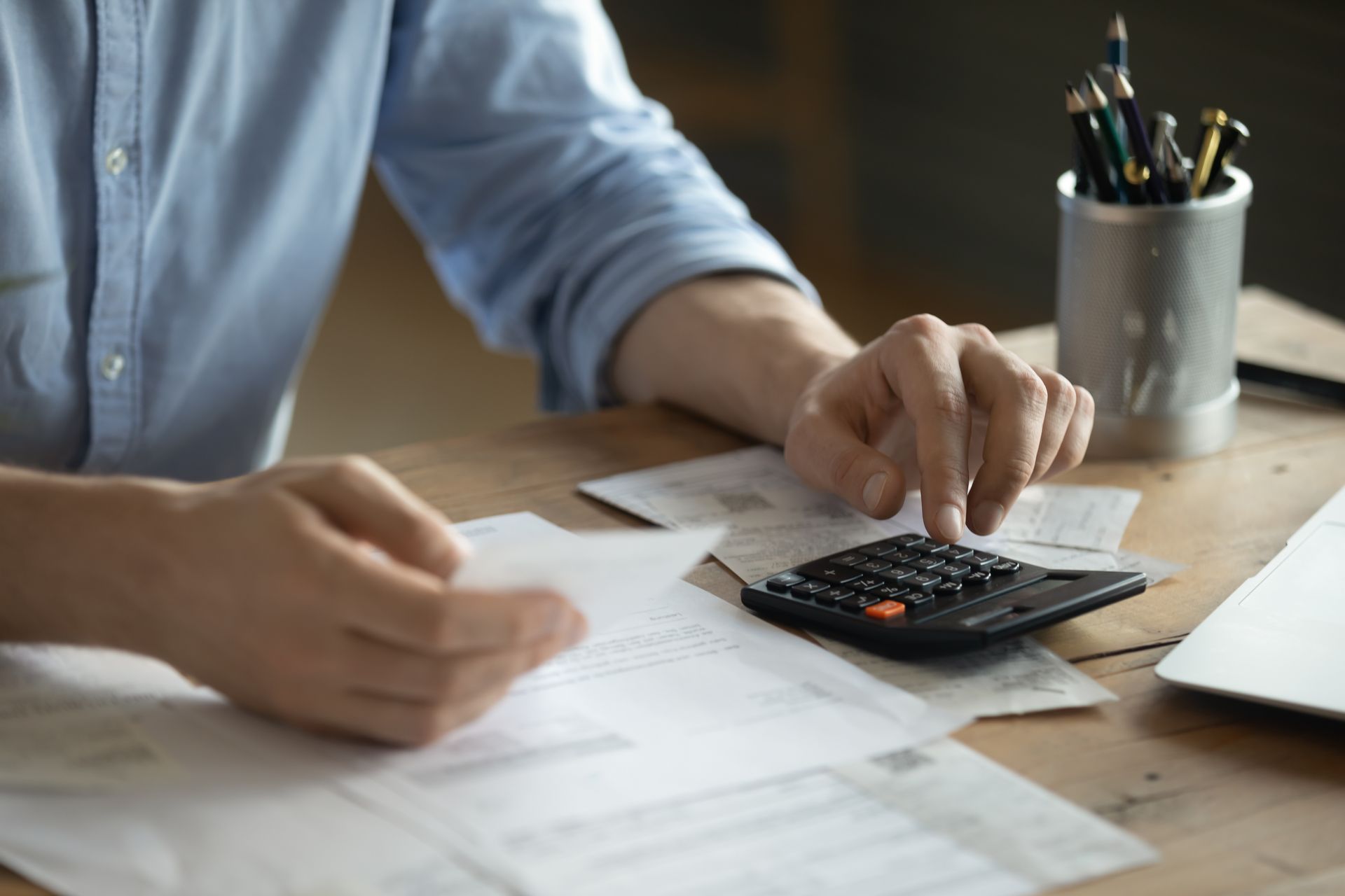 Hands using a calculator and paperwork on a wooden desk beside a laptop and pencil cup Hands using a calculator and paperwork on a wooden desk beside a laptop and pencil cup