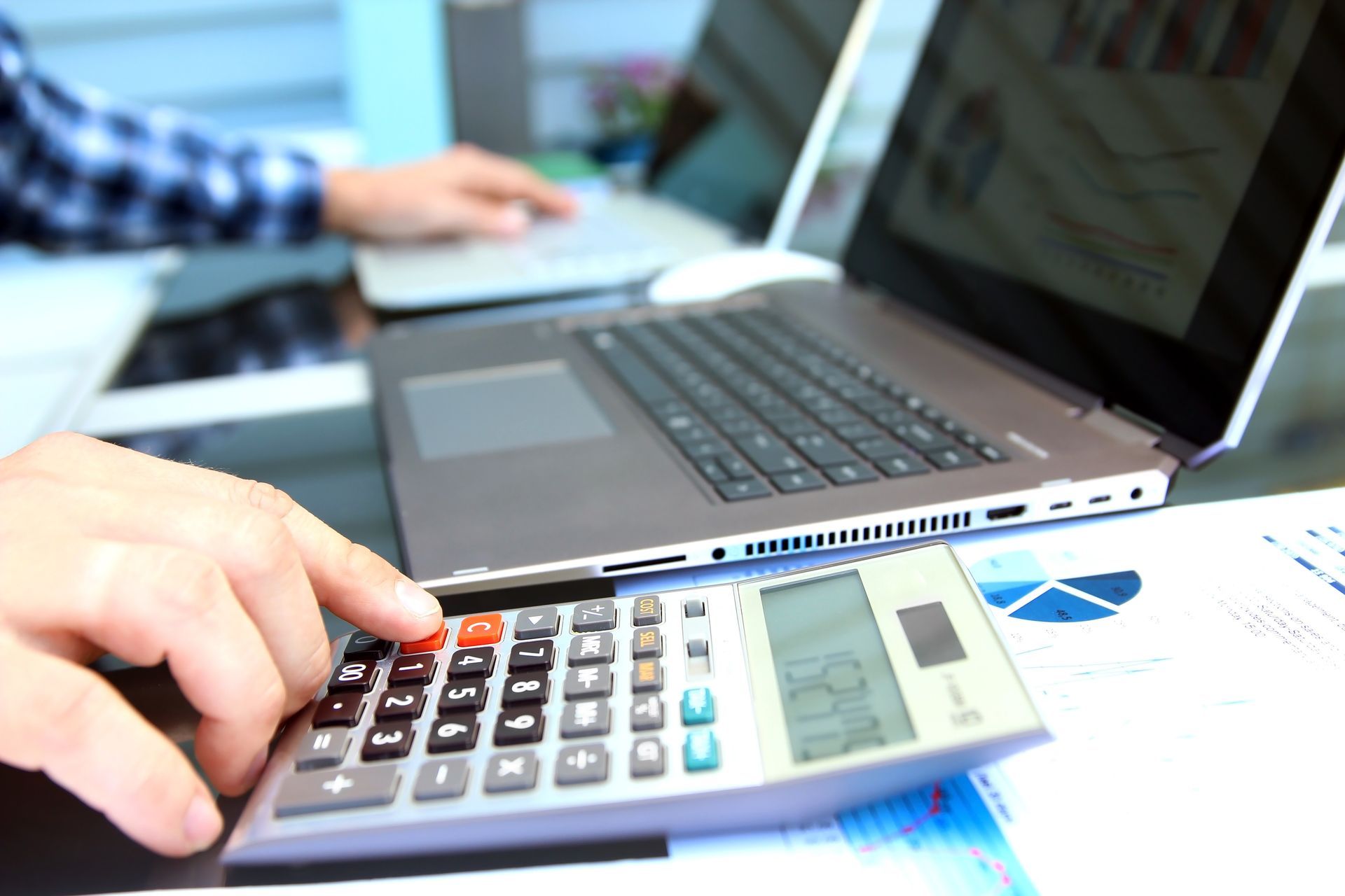 Hand using calculator beside laptop and business charts on a desk Hand using calculator beside laptop and business charts on a desk
