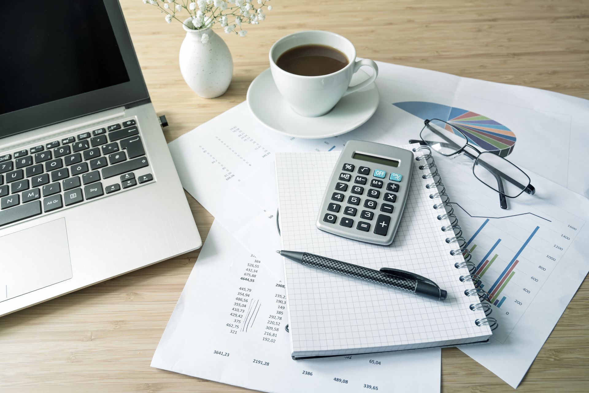 Laptop, coffee cup, calculator, and papers on a wooden desk, possibly for financial analysis. Laptop, coffee cup, calculator, and papers on a wooden desk, possibly for financial analysis.