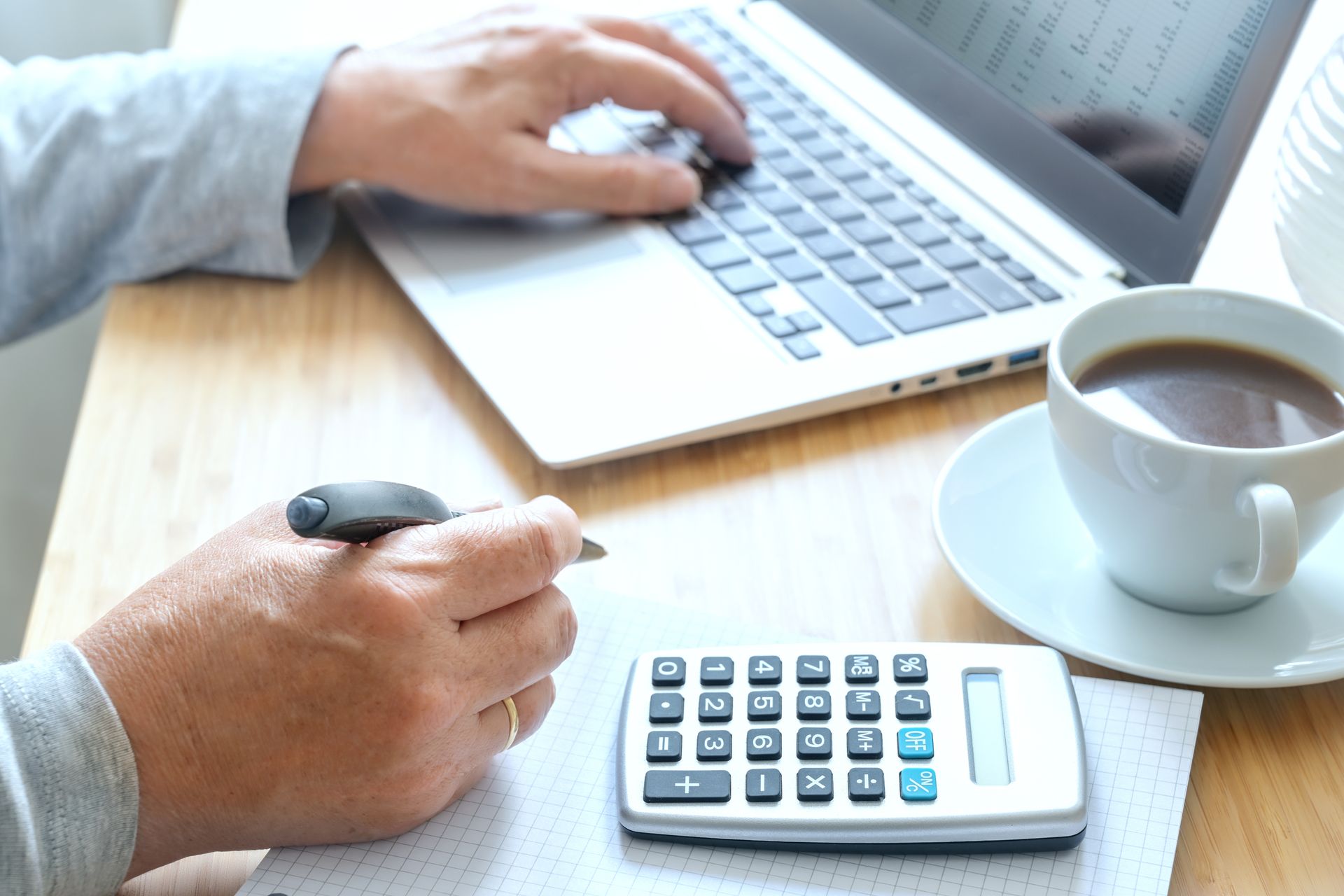 Person typing on laptop, writing notes, and using a calculator next to a cup of coffee. Person typing on laptop, writing notes, and using a calculator next to a cup of coffee.