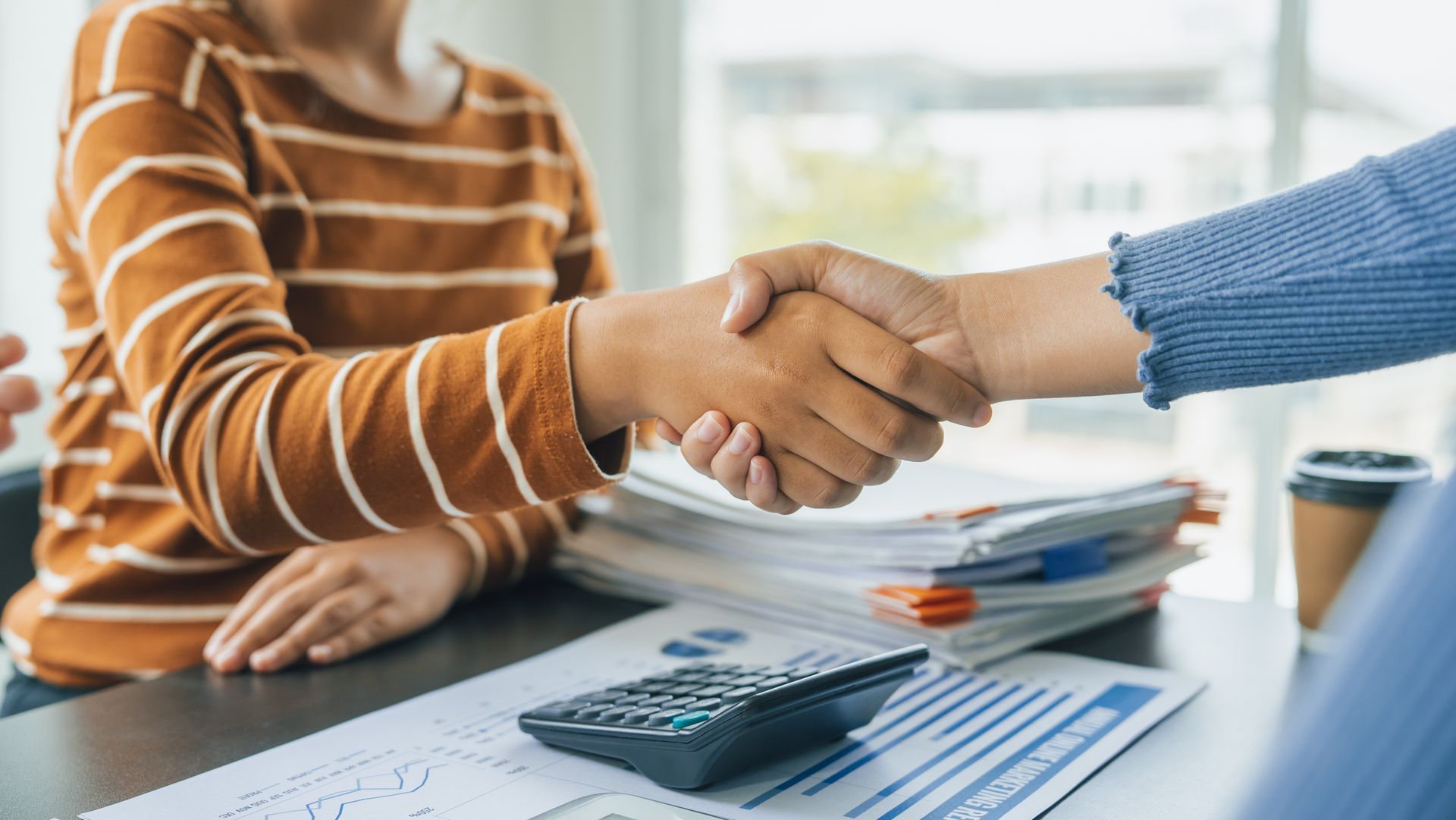 Two people shake hands over a desk with a calculator, documents, and a coffee cup, representing a business agreement. Two people shake hands over a desk with a calculator, documents, and a coffee cup, representing a business agreement.