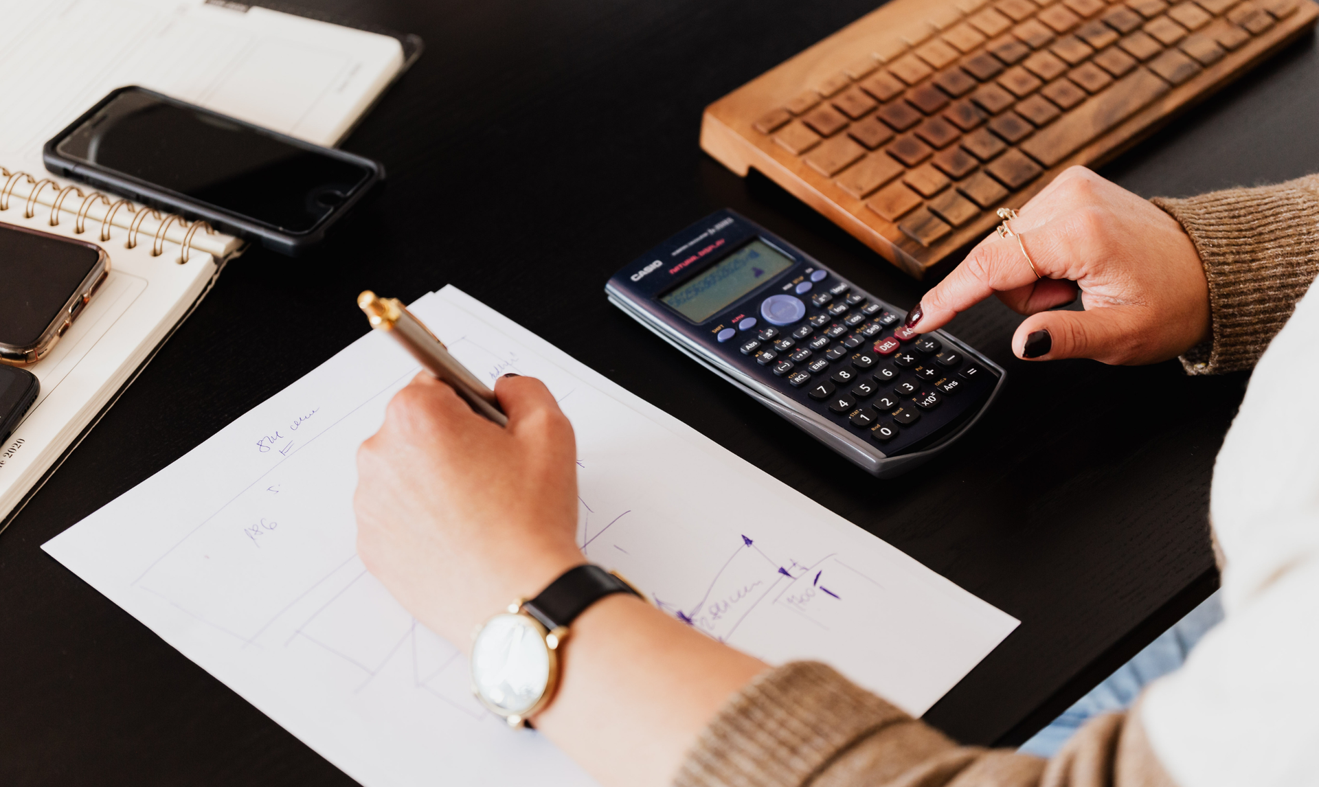 Person using a calculator and pen, writing on paper at a desk. A phone, keyboard, and notebook are also present. Person using a calculator and pen, writing on paper at a desk. A phone, keyboard, and notebook are also present.