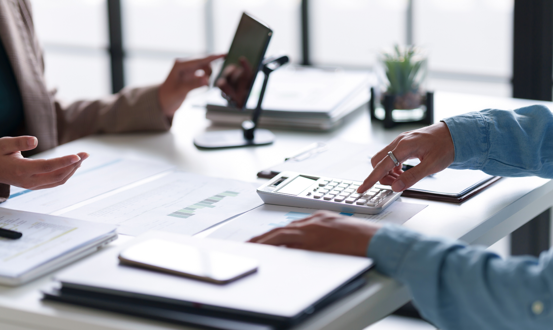 Two people at a desk, reviewing documents and using a calculator. A tablet and a smartphone are also present. Two people at a desk, reviewing documents and using a calculator. A tablet and a smartphone are also present.