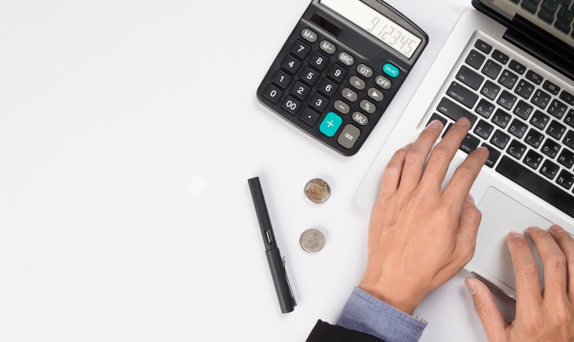 Hands typing on a laptop with a calculator, pen, and coins on a white surface. Hands typing on a laptop with a calculator, pen, and coins on a white surface.