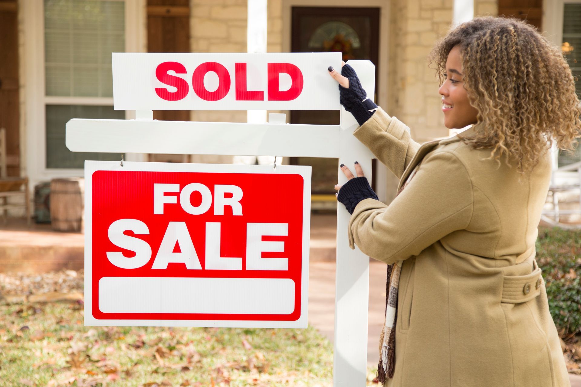 A woman stands in front of a home with a 'Sold' sign, showcasing a local realtor company's services.