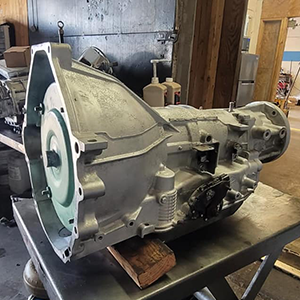 Transmission housing on a workbench in a garage, shown in dusty gray metal with attached components.