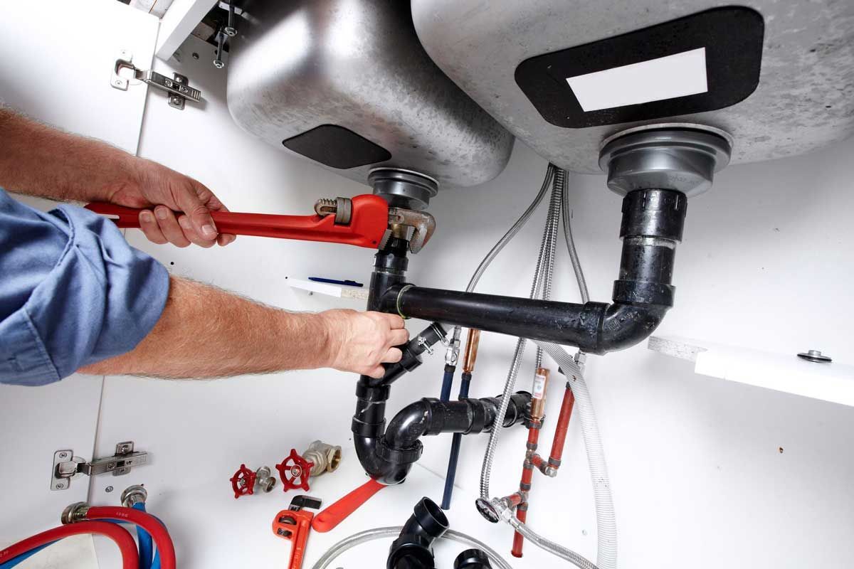 Plumber tightening pipe connections under a double kitchen sink using a red wrench.
