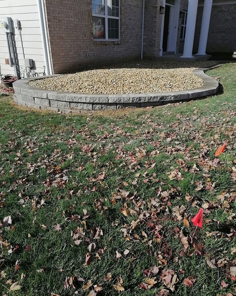 Raised stone garden bed beside a brick house, filled with gravel and surrounded by lawn and fallen leaves.