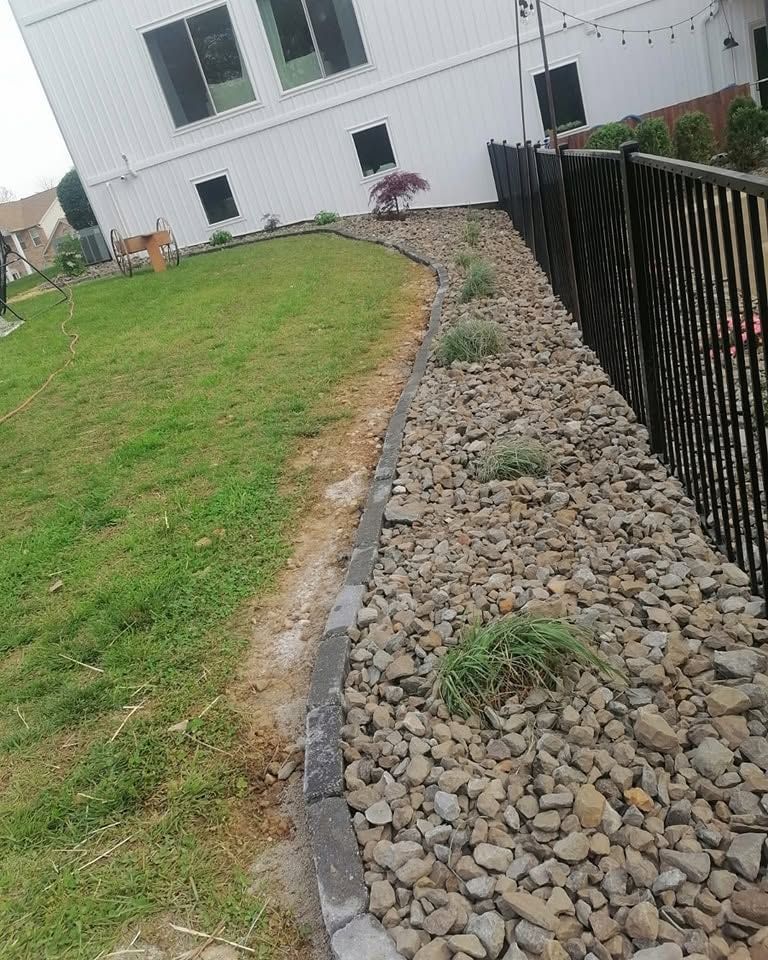 Rock-lined drainage path beside a green lawn and black metal fence next to a white house.