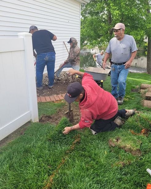 Three people working in a yard, one kneeling in the grass near a fence and others standing by a house.
