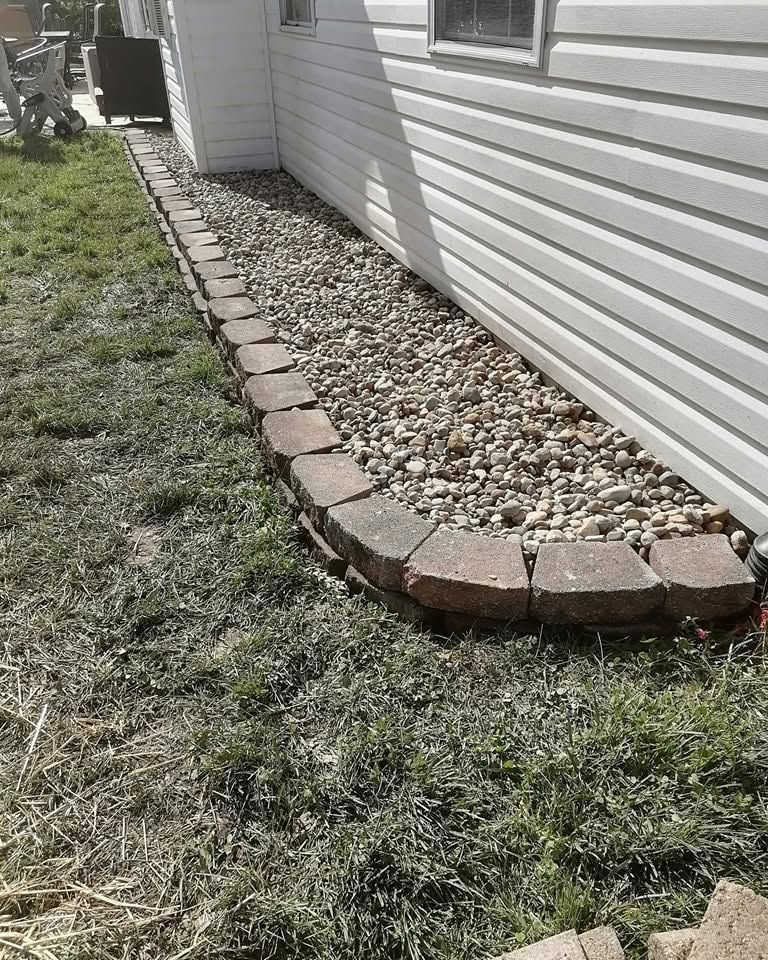 Curved stone border around white house siding with decorative rocks and grass edging