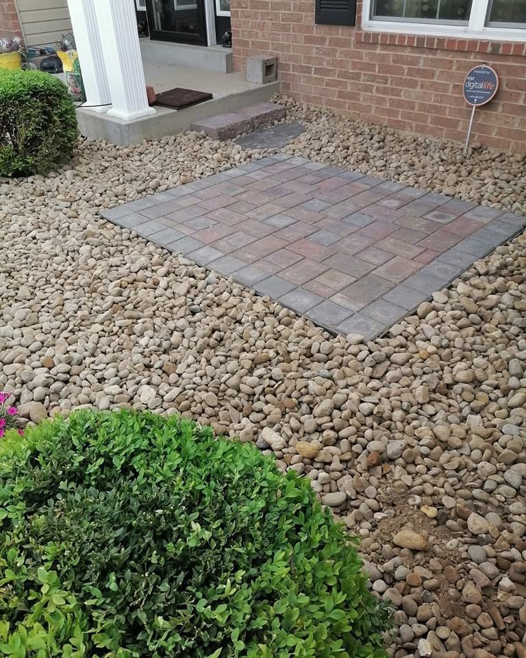 Front porch with a small brick paver patio surrounded by decorative river rocks and shrubs