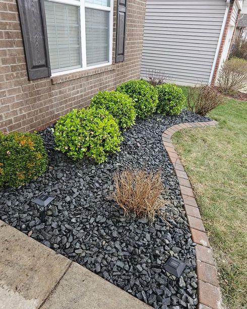 Front yard landscaping with black rock mulch, green shrubs, dry grass, and brick edging beside a house