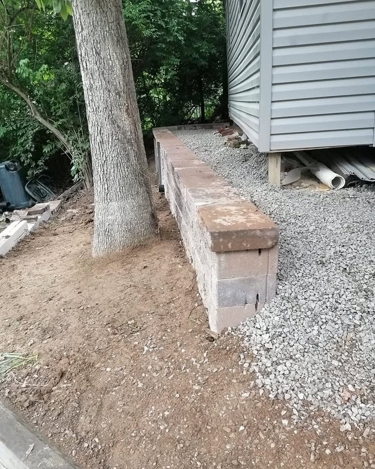 Dirt and gravel beside a house, with a large tree and a low concrete retaining ledge along the wall.