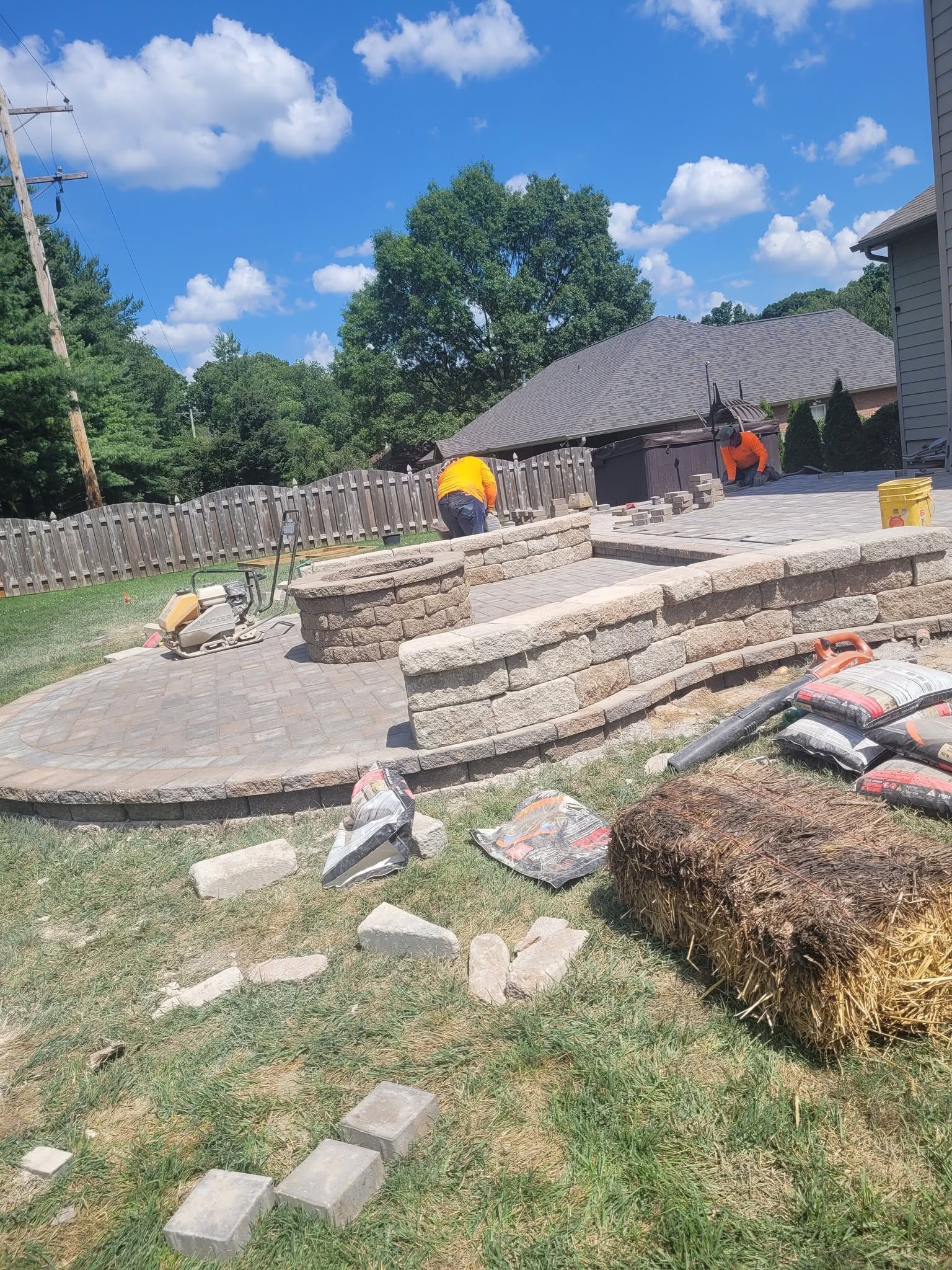 Stone patio and low retaining walls under construction in a sunny backyard, with workers and scattered materials