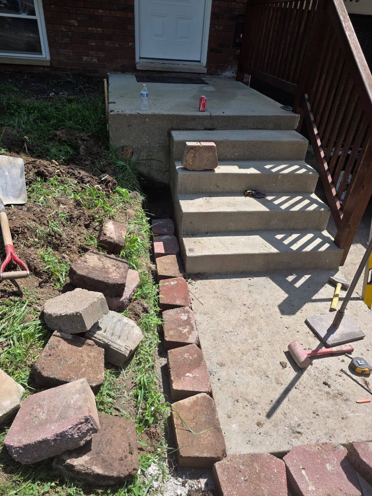Front steps by a porch, with loose bricks and a shovel beside a concrete walkway.