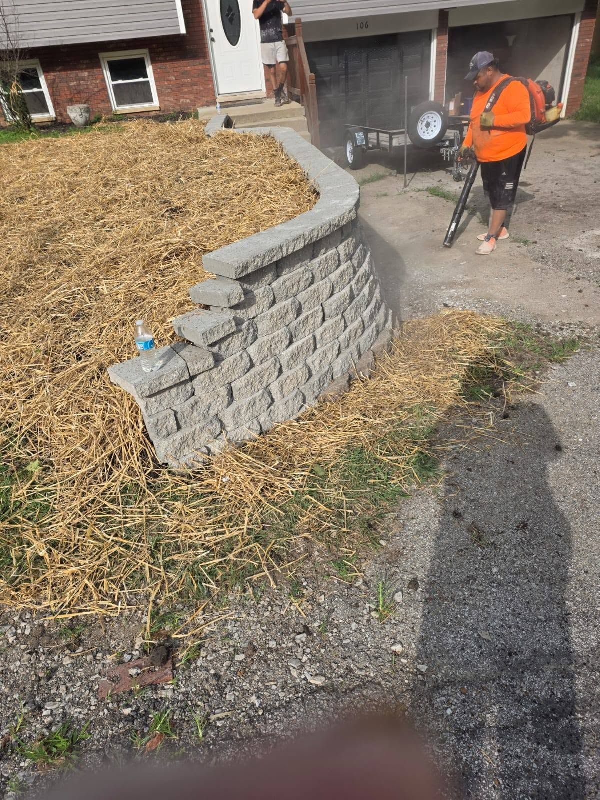 Worker in orange vest using a power tool near a stone retaining wall and wood mulch at a house