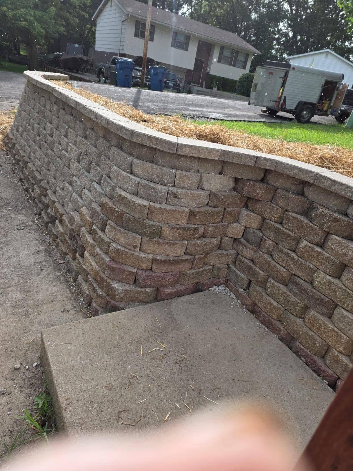 Curved stone retaining wall beside a driveway, with a house and parked trailer in the background.