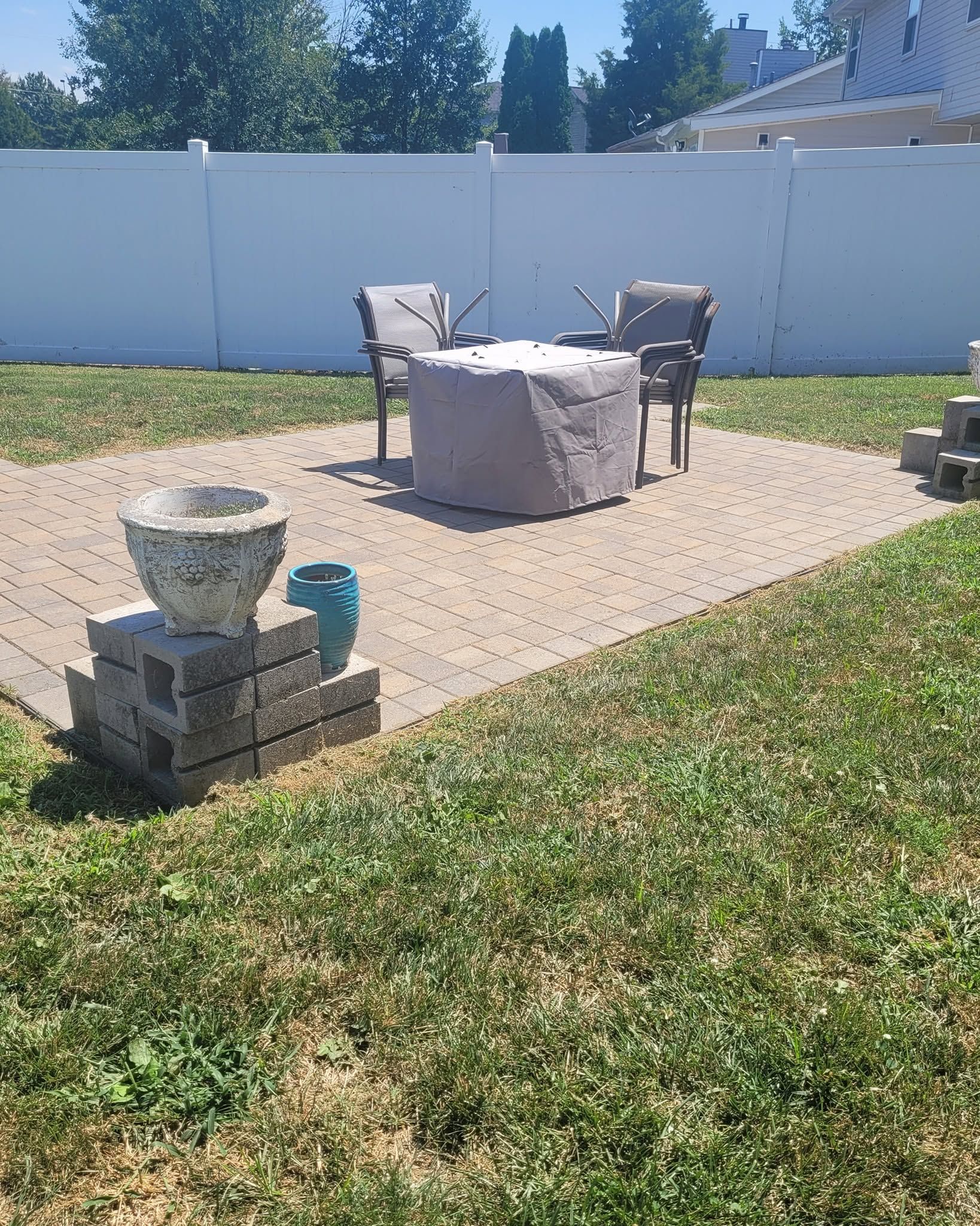 Backyard patio with stone seating, covered table, and white privacy fence under bright sun