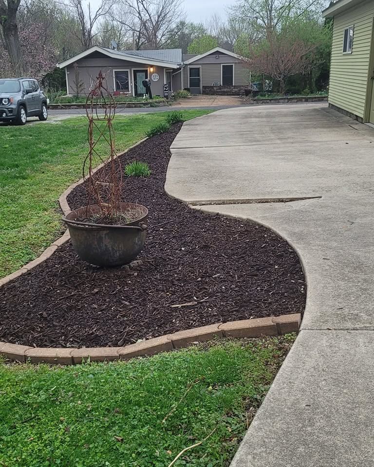 Curved concrete driveway beside a mulched planting bed with a potted sapling and grassy lawn