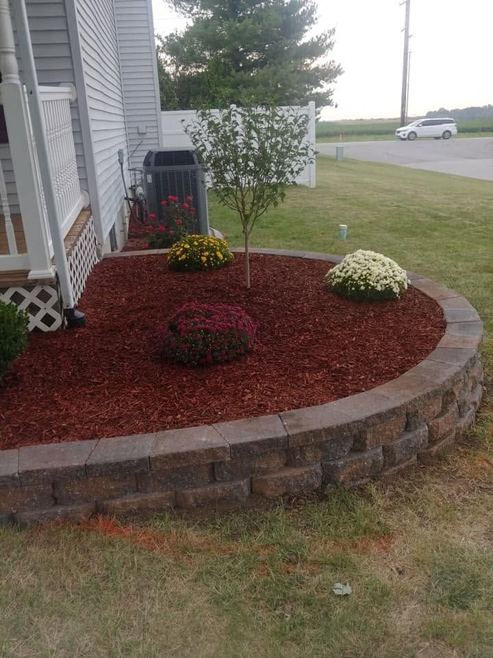 Curved landscaped flower bed with red mulch, white and yellow flowers, and a small tree beside a house.