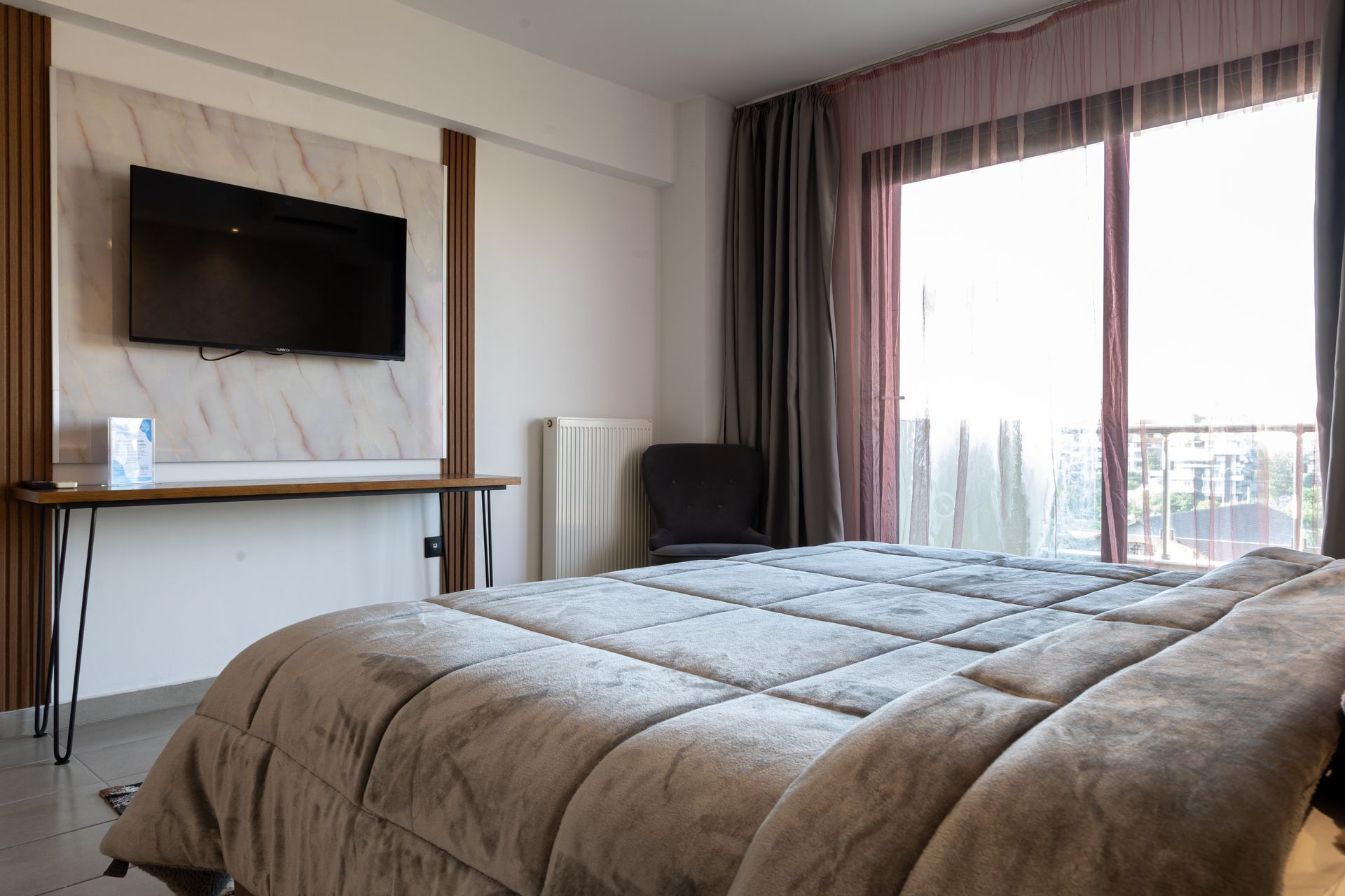 Modern bedroom with a bed, wall-mounted TV, desk, and large window with balcony view.