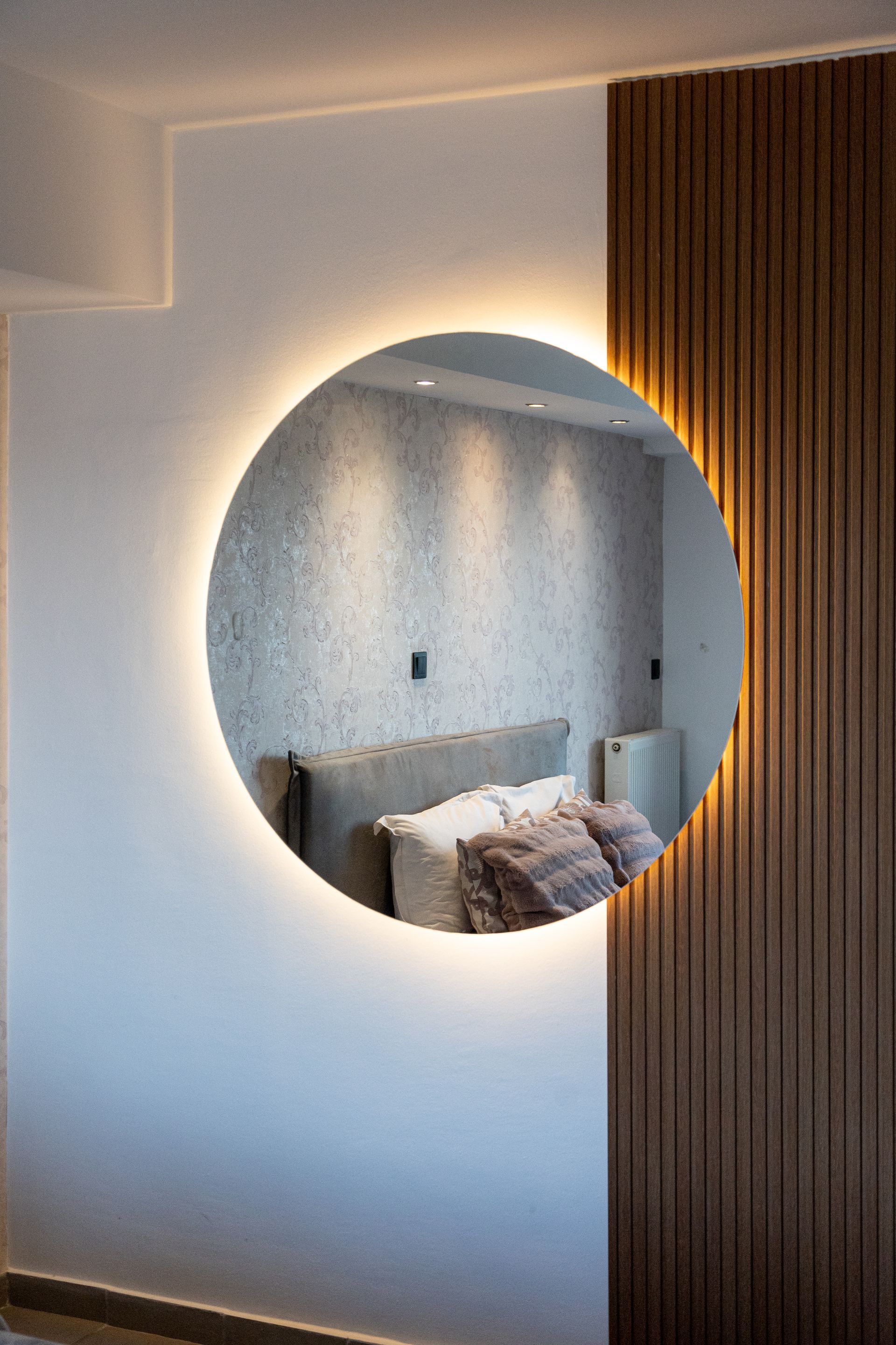 Backlit round wall mirror reflecting a cozy bedroom with gray headboard and pillows, beside wood paneling