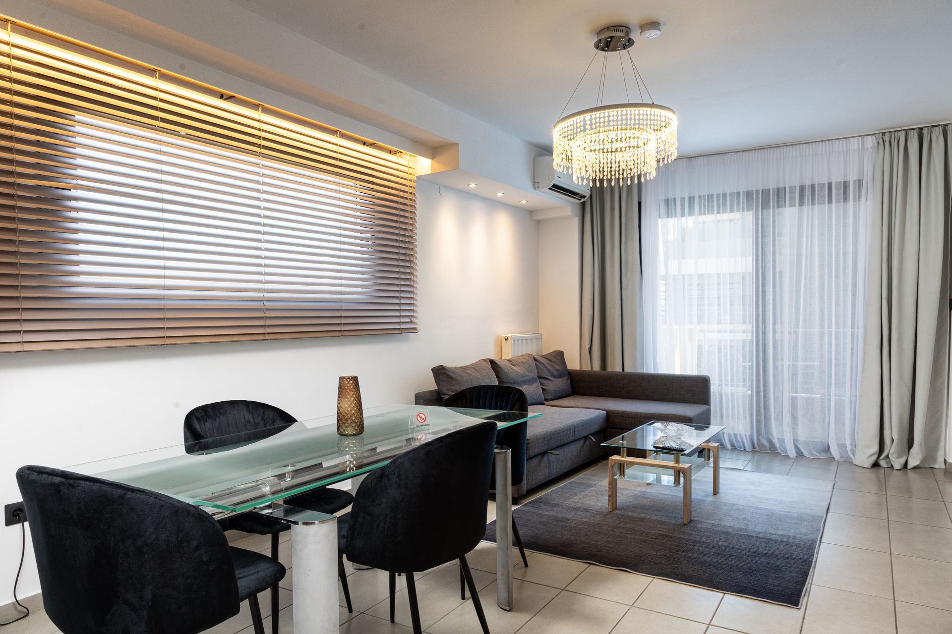 Bright modern living-dining room with a sofa, glass table, black chairs, and large windows with blinds and curtains